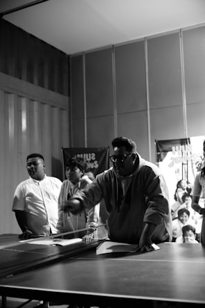 A group of people gather around a ping pong table indoors, with one person appearing to be in action, possibly making a move in a game. The scene is captured in black and white, and the background features a banner and an audience observing the activity.