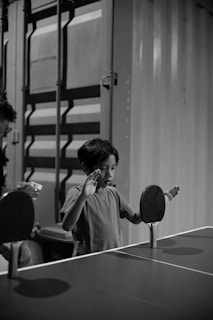 A black and white photo of a child standing behind a table with two vertically mounted table tennis paddles. The child is wearing a plain t-shirt and is looking at the paddles with an expression of curiosity or concentration. In the background, there is a large corrugated metal surface, possibly a shipping container.