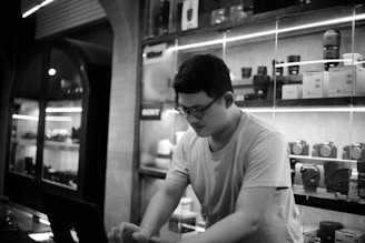 A person wearing glasses and a t-shirt stands in front of illuminated shelves filled with camera lenses and boxes in a store. The scene is in black and white, giving it a classic feel. The person appears to be focused on something below them, possibly a laptop.