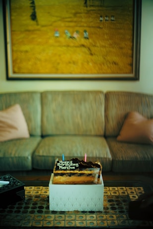 A birthday cake placed on a decorative table with geometric patterns, set in front of a sofa with two cushions. Above the sofa hangs a large painting depicting a field scene. The cake is topped with chocolate and two candles, accompanied by a birthday message.