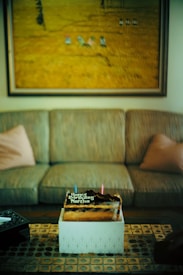 A birthday cake placed on a decorative table with geometric patterns, set in front of a sofa with two cushions. Above the sofa hangs a large painting depicting a field scene. The cake is topped with chocolate and two candles, accompanied by a birthday message.