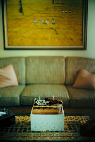A birthday cake placed on a decorative table with geometric patterns, set in front of a sofa with two cushions. Above the sofa hangs a large painting depicting a field scene. The cake is topped with chocolate and two candles, accompanied by a birthday message.