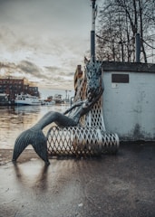 A mermaid sculpture made of metal is leaning against a wire mesh bench on a waterfront walkway. In the background, there are boats docked on a calm body of water, with buildings and a construction crane visible. The sky is overcast with dense clouds, and leafless trees line the shore.