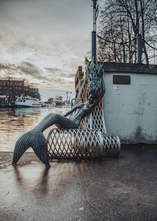A mermaid sculpture made of metal is leaning against a wire mesh bench on a waterfront walkway. In the background, there are boats docked on a calm body of water, with buildings and a construction crane visible. The sky is overcast with dense clouds, and leafless trees line the shore.