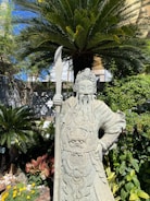 An intricately carved stone statue of a warrior figure holding a spear stands amidst lush greenery. The figure is adorned with elaborate armor and a detailed headpiece. Behind the statue, a large palm tree fans out its leaves, complementing the dense foliage surrounding it. Bright sunlight casts shadows across the scene, highlighting the textures of the stone and the vibrant hues of the plants.