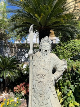 An intricately carved stone statue of a warrior figure holding a spear stands amidst lush greenery. The figure is adorned with elaborate armor and a detailed headpiece. Behind the statue, a large palm tree fans out its leaves, complementing the dense foliage surrounding it. Bright sunlight casts shadows across the scene, highlighting the textures of the stone and the vibrant hues of the plants.