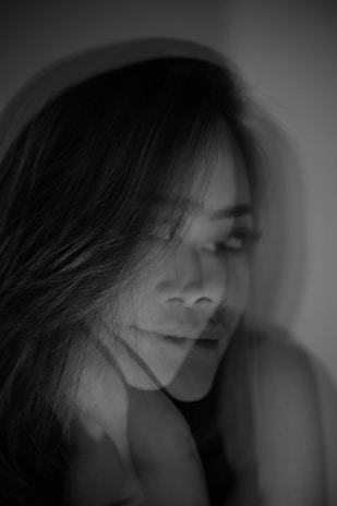A monochrome photograph featuring a woman's face in motion, creating a double-exposure effect. Her eyes are closed, and her lips show a subtle, serene expression. The interplay of light and shadow adds a dreamlike quality to the image.