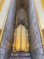 An intricately decorated golden door with floral patterns, framed by golden and marble walls. The door features a pointed arch and a small oval window, indicating it might be part of a temple or similar structure.
