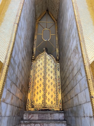 An intricately decorated golden door with floral patterns, framed by golden and marble walls. The door features a pointed arch and a small oval window, indicating it might be part of a temple or similar structure.