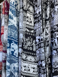 An array of patterned textile fabrics in various colors, including maroon, blue, and black, featuring intricate white designs. The motifs are predominantly elephants and ornate floral patterns.