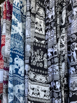 An array of patterned textile fabrics in various colors, including maroon, blue, and black, featuring intricate white designs. The motifs are predominantly elephants and ornate floral patterns.