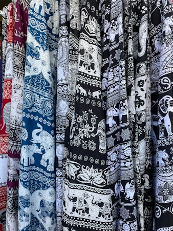 An array of patterned textile fabrics in various colors, including maroon, blue, and black, featuring intricate white designs. The motifs are predominantly elephants and ornate floral patterns.