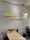 A minimalist interior with a white brick wall displaying neon yellow Thai script. The room features a small wooden table set against the wall, equipped with condiments and utensils. Black metal chairs surround the table, and a single wall lamp provides soft lighting.