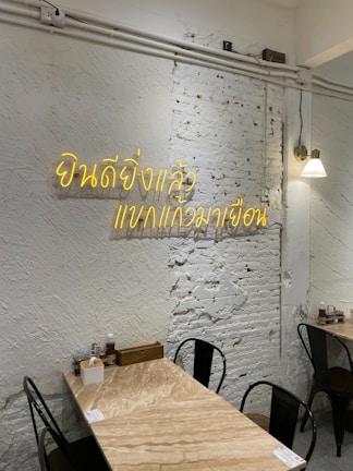 A minimalist interior with a white brick wall displaying neon yellow Thai script. The room features a small wooden table set against the wall, equipped with condiments and utensils. Black metal chairs surround the table, and a single wall lamp provides soft lighting.