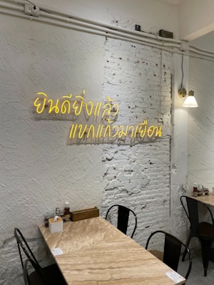 A minimalist interior with a white brick wall displaying neon yellow Thai script. The room features a small wooden table set against the wall, equipped with condiments and utensils. Black metal chairs surround the table, and a single wall lamp provides soft lighting.