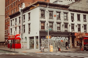 A city street corner featuring an old building with graffiti-covered lower walls and a vibrant red taco restaurant sign. There is a pedestrian walking by, a crosswalk, and a pole with street signs. Autumnal trees are on the sidewalk, and the overall scene is urban and gritty.