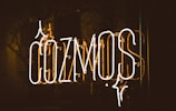 Neon sign displaying the word 'COZMOS' with a dark background. The letters are illuminated in a warm, glowing light, and there are reflections visible on a glass surface.