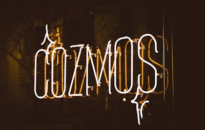 Neon sign displaying the word 'COZMOS' with a dark background. The letters are illuminated in a warm, glowing light, and there are reflections visible on a glass surface.