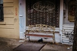 An ornate bench with a metal vine design stands in front of a shuttered shop with graffiti on the walls. The shop sign reads 'Psychic' and the walls are painted yellow. A small cartoon figure and scribbles are visible on the left wall.