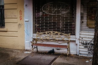 An ornate bench with a metal vine design stands in front of a shuttered shop with graffiti on the walls. The shop sign reads 'Psychic' and the walls are painted yellow. A small cartoon figure and scribbles are visible on the left wall.
