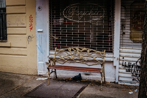 An ornate bench with a metal vine design stands in front of a shuttered shop with graffiti on the walls. The shop sign reads 'Psychic' and the walls are painted yellow. A small cartoon figure and scribbles are visible on the left wall.