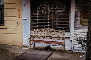 An ornate bench with a metal vine design stands in front of a shuttered shop with graffiti on the walls. The shop sign reads 'Psychic' and the walls are painted yellow. A small cartoon figure and scribbles are visible on the left wall.