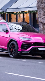 A bright pink luxury SUV is parked on the side of the street. The vehicle features sleek, angular lines and large black rims. In the background, there is a building with festive greenery decorations and a sign reading 'High Road House Brasserie'. A person is visible near the vehicle, though not clearly identifiable.