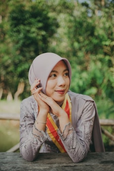 A person wearing a light-colored hijab and a decorative dress decorated with intricate patterns appears content while resting their head gently on their hands. They are sitting at an outdoor table with lush green foliage in the background, creating a serene atmosphere.