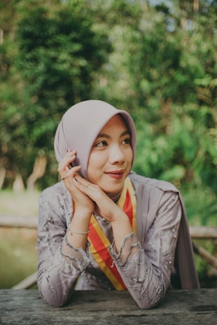 A person wearing a light-colored hijab and a decorative dress decorated with intricate patterns appears content while resting their head gently on their hands. They are sitting at an outdoor table with lush green foliage in the background, creating a serene atmosphere.