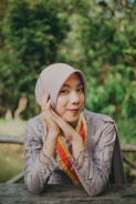 A woman wearing a light purple hijab and a detailed, embroidered dress with a red and yellow sash smiles gently with her hands resting under her chin. The setting is outdoors, with a blurred background full of lush green foliage.