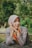 A woman wearing a light purple hijab and a detailed, embroidered dress with a red and yellow sash smiles gently with her hands resting under her chin. The setting is outdoors, with a blurred background full of lush green foliage.