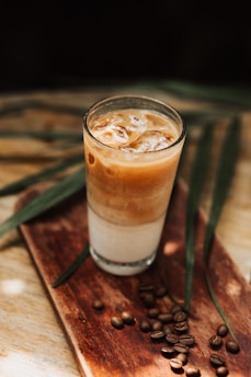 A glass of iced coffee sits on a wooden board, surrounded by coffee beans and green leaves. The drink has layers, with a creamy bottom and a darker coffee layer on top.
