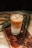 A glass of iced coffee sits on a wooden board, surrounded by coffee beans and green leaves. The drink has layers, with a creamy bottom and a darker coffee layer on top.