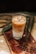 A glass of iced coffee sits on a wooden board, surrounded by coffee beans and green leaves. The drink has layers, with a creamy bottom and a darker coffee layer on top.