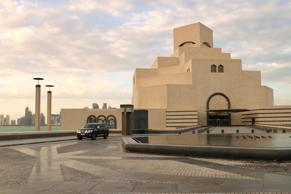 A modern architectural building with stacked geometric layers made from light-colored stone sits beside a reflective body of water. A black SUV is parked on a stone-paved area nearby. In the background, city skyscrapers rise along a distant skyline against a sky filled with soft, scattered clouds.