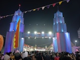 A large crowd of people gathers at a festival beneath a decorative entrance resembling towers with vibrant blue and red lighting. The area is adorned with colorful flags and banners, creating a festive atmosphere.