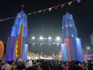 A large crowd of people gathers at a festival beneath a decorative entrance resembling towers with vibrant blue and red lighting. The area is adorned with colorful flags and banners, creating a festive atmosphere.