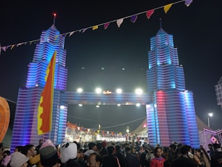 A large crowd of people gathers at a festival beneath a decorative entrance resembling towers with vibrant blue and red lighting. The area is adorned with colorful flags and banners, creating a festive atmosphere.