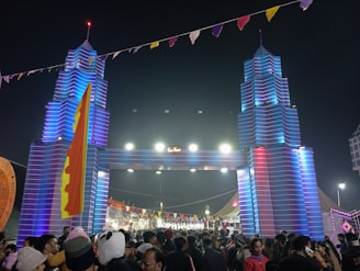 A large crowd of people gathers at a festival beneath a decorative entrance resembling towers with vibrant blue and red lighting. The area is adorned with colorful flags and banners, creating a festive atmosphere.