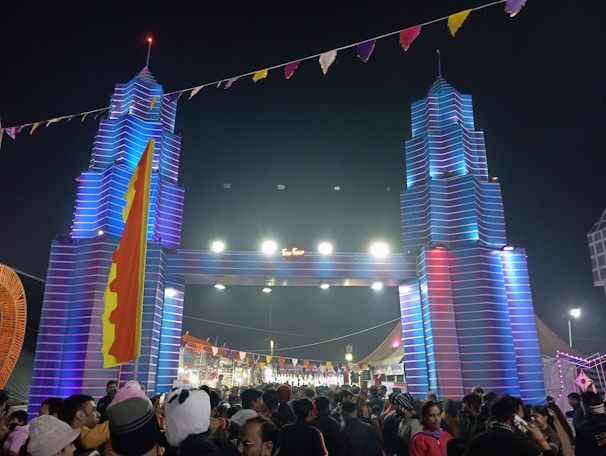 A large crowd of people gathers at a festival beneath a decorative entrance resembling towers with vibrant blue and red lighting. The area is adorned with colorful flags and banners, creating a festive atmosphere.