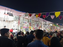 A vibrant market stall adorned with a variety of hanging kitchen utensils, including ladles and spatulas, alongside colorful decorations and jewelry items. The stall is busy, with a crowd of people browsing and engaging with the goods on display. Brightly colored pennant banners hang overhead, adding to the festive atmosphere.