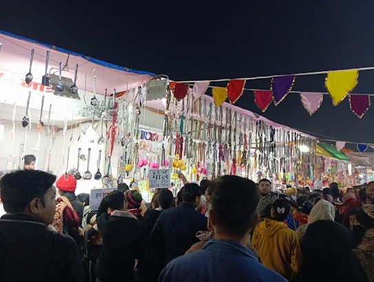 A vibrant market stall adorned with a variety of hanging kitchen utensils, including ladles and spatulas, alongside colorful decorations and jewelry items. The stall is busy, with a crowd of people browsing and engaging with the goods on display. Brightly colored pennant banners hang overhead, adding to the festive atmosphere.