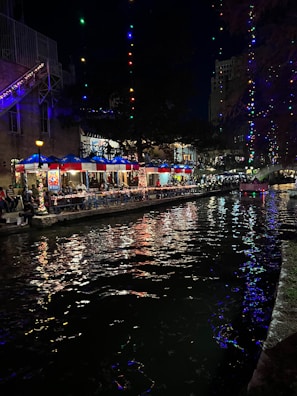 A lively scene at night featuring a river bordered by illuminated walkways and a lineup of restaurants. Colorful lights reflect on the water, adding vibrancy. The area is bustling with people dining under blue canopies adorned with patterns resembling the Texas flag.