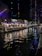 A lively scene at night featuring a river bordered by illuminated walkways and a lineup of restaurants. Colorful lights reflect on the water, adding vibrancy. The area is bustling with people dining under blue canopies adorned with patterns resembling the Texas flag.