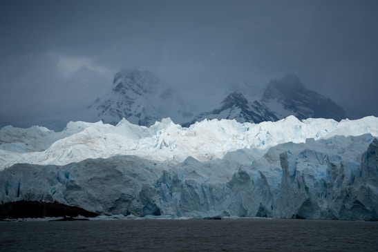 A vast, rugged glacier extends across the foreground, with sharp, jagged ice formations and a mix of bright white and pale blue hues. Looming in the background are tall, dark mountains partially obscured by thick, gray clouds, creating a dramatic contrast with the icy landscape below.