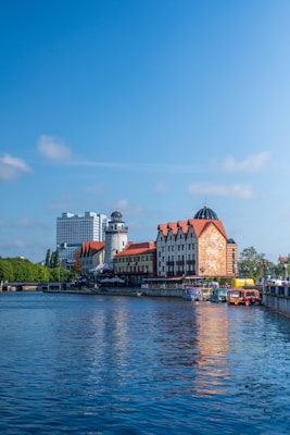 A picturesque waterfront scene features buildings with traditional architecture, including a tower with a dome roof. The structures have a mix of modern and classic designs, with one facade displaying an artistic mural. Boats are docked along the river, and the sky is clear with a few scattered clouds.