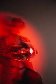 A blurred and abstract image featuring a person partially obscured in red lighting. They appear to be holding a reflective disco ball, with light reflections creating an intense red hue across the scene. The overall atmosphere is atmospheric and moody.