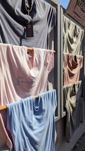 Several t-shirts are displayed on a rack outdoors, attached to wooden rods that fit into a wooden frame. The shirts vary in color including light blue, pink, and gray, and feature simple designs or text with 'Santorini, Greece' visible on some. Sunlight casts shadows across the shirts, suggesting a sunny day.