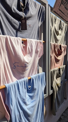 Several t-shirts are displayed on a rack outdoors, attached to wooden rods that fit into a wooden frame. The shirts vary in color including light blue, pink, and gray, and feature simple designs or text with 'Santorini, Greece' visible on some. Sunlight casts shadows across the shirts, suggesting a sunny day.