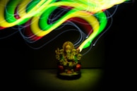 A colorful, glowing light painting swirls around a small statue of a deity with an elephant head, which is seated and adorned with intricate details. The lights create a dynamic and energetic pattern above the statue, illuminating it from above and highlighting its features.